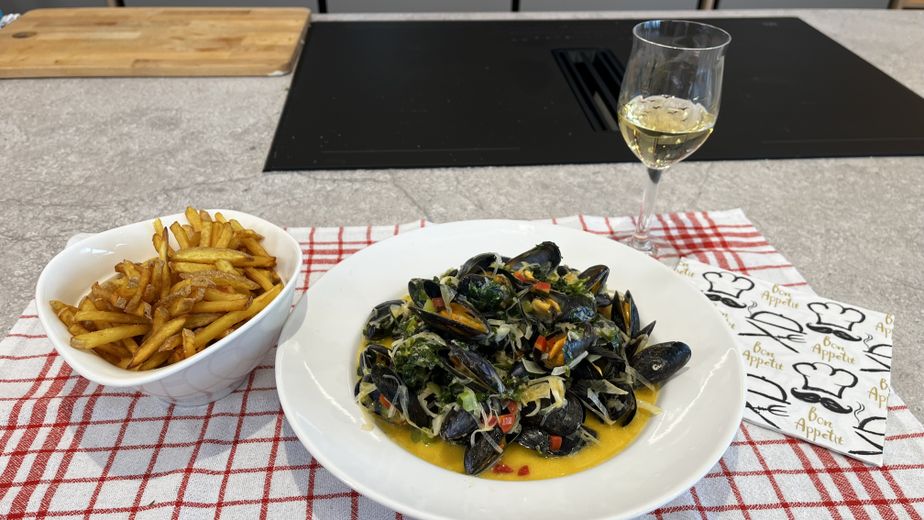 Ein fertig angerichteter Teller Moules Frites in einer hellen Sauce mit Kräutern und Gemüse, serviert mit einer Schüssel goldbraun frittierter Pommes Frites und einem Glas Weißwein. Das Ensemble ist auf einem rot-weiß karierten Tuch in einer modernen Küche vor einem schwarzen Induktionskochfeld präsentiert.