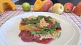Ein weißer Teller mit fein aufgeschnittenem Rinder-Carpaccio auf einer cremigen, orangefarbenen Kürbiscreme. Das Gericht ist garniert mit knusprigen Parmesanchips, frischen grünen Kräutern und etwas Chili-Öl. Im leicht verschwommenen Hintergrund liegen herbstliche Zutaten wie Kürbisse und Äpfel auf einem rot-weiß karierten Tuch.