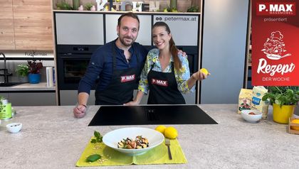 Max Impertro und Tanja stehen lächelnd nebeneinander in einer modernen Küche hinter einer Kochinsel. Im Vordergrund ist ein fertig angerichteter Teller mit Tortellini und einer Garnele auf einem hellgrünen Platzset zu sehen. Tanja hält eine Zitrone in der Hand, während im Hintergrund Backzutaten bereitstehen.