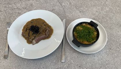 Das fertig angerichtete Rib-Eye-Steak auf einem weißen Teller, aufgeschnitten und mit der Pfeffersauce und dem Zwiebelkompott bedeckt. Daneben steht in einer kleinen schwarzen Schale das goldbraun gebackene Erdäpfel-Gratin, das reichlich mit Schnittlauch bestreut ist.