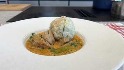 Servierfertiges Kalbsrahmgulasch in cremiger Sauce mit zarten Fleischstücken und einem großen Semmelknödel auf einem weißen Teller. Das Gericht ist mit frischer Petersilie und einer Essiggurke garniert.