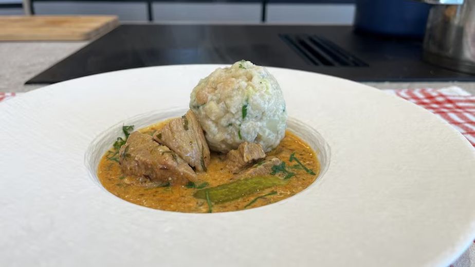 Servierfertiges Kalbsrahmgulasch in cremiger Sauce mit zarten Fleischstücken und einem großen Semmelknödel auf einem weißen Teller. Das Gericht ist mit frischer Petersilie und einer Essiggurke garniert.