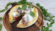 Die fertige Pizza Bianca liegt auf einem dunklen Holzbrett, umgeben von frischem Rucola und Basilikum. Ein Stück ist herausgeschnitten und zeigt den Belag aus Pistazienpesto, Prosciutto, Mozzarella und marinierten Tomaten, großzügig bestreut mit geriebenem Parmesan.