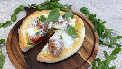Die fertige Pizza Bianca liegt auf einem dunklen Holzbrett, umgeben von frischem Rucola und Basilikum. Ein Stück ist herausgeschnitten und zeigt den Belag aus Pistazienpesto, Prosciutto, Mozzarella und marinierten Tomaten, großzügig bestreut mit geriebenem Parmesan.
