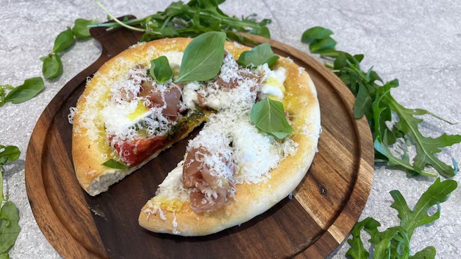 Die fertige Pizza Bianca liegt auf einem dunklen Holzbrett, umgeben von frischem Rucola und Basilikum. Ein Stück ist herausgeschnitten und zeigt den Belag aus Pistazienpesto, Prosciutto, Mozzarella und marinierten Tomaten, großzügig bestreut mit geriebenem Parmesan.
