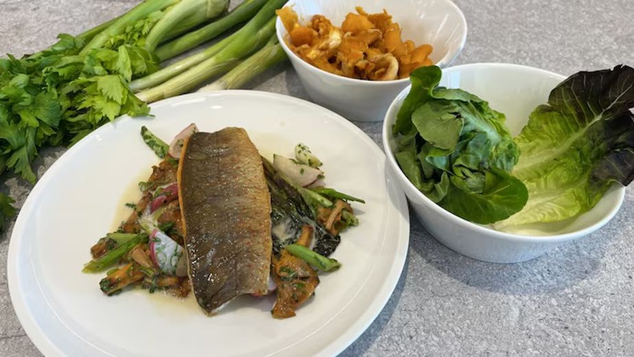 Fertig angerichteter Teller mit gebratenem Saibling auf Gemüse und Sherry-Sauce. Im Hintergrund sind einige der frischen Zutaten für das Rezept zu sehen: Pilze, Romanasalat und Stangensellerie.