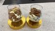 Zwei Gläser gefüllt mit geschichtetem Apfel-Crumble, bestehend aus heller Frischkäsecreme, goldbraun gebackenen Streuseln und gedünsteten Apfelstücken. Die Desserts sind mit feiner grüner Zitruszeste garniert und ansprechend auf runden, goldfarbenen Podesten angerichtet.
