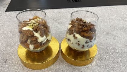 Zwei Gläser gefüllt mit geschichtetem Apfel-Crumble, bestehend aus heller Frischkäsecreme, goldbraun gebackenen Streuseln und gedünsteten Apfelstücken. Die Desserts sind mit feiner grüner Zitruszeste garniert und ansprechend auf runden, goldfarbenen Podesten angerichtet.