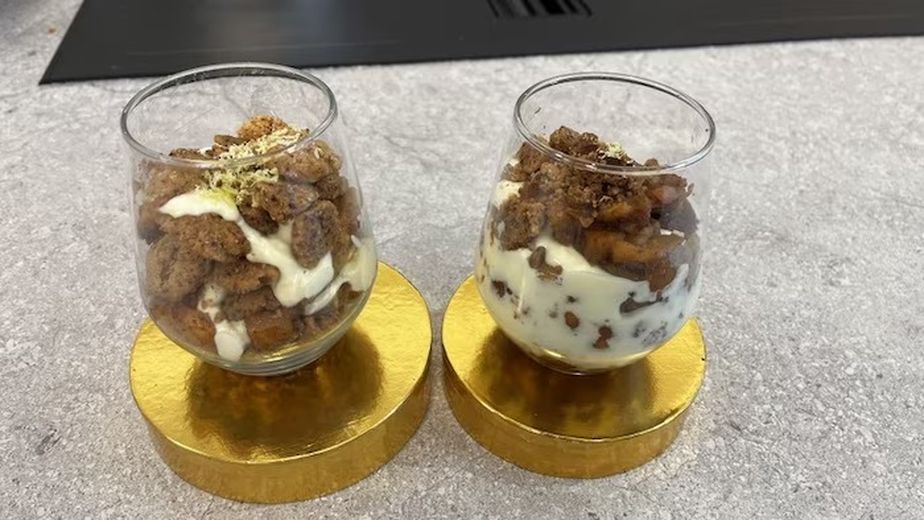 Zwei Gläser gefüllt mit geschichtetem Apfel-Crumble, bestehend aus heller Frischkäsecreme, goldbraun gebackenen Streuseln und gedünsteten Apfelstücken. Die Desserts sind mit feiner grüner Zitruszeste garniert und ansprechend auf runden, goldfarbenen Podesten angerichtet.