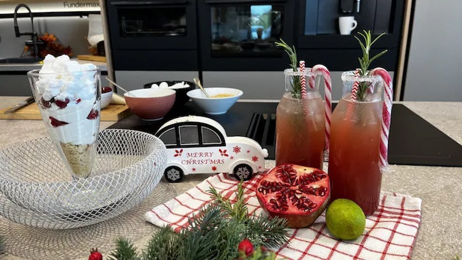 Ein festlich angerichtetes Schneeflocken-Dessert in einem hohen Glas mit sichtbaren Schichten aus Kekskrümeln, Himbeeren und weißer Creme, getoppt mit Baiser. Daneben stehen zwei rötliche Mocktails mit Zuckerrand, Rosmarinzweigen und Zuckerstangen, dekoriert mit einem halbierten Granatapfel und weihnachtlichen Accessoires auf einer modernen Küchenarbeitsplatte.