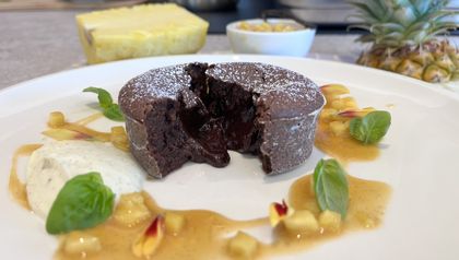 Ein kleiner, mit Puderzucker bestäubter Schokokuchen, der in der Mitte aufgeschnitten ist, sodass der dunkle, flüssige Kern herausläuft. Der Kuchen ist dekorativ auf einem weißen Teller zusammen mit einer hellen Mascarpone-Creme, frischen Basilikumblättern und einer Ananas-Karamell-Sauce mit kleinen Fruchtstücken angerichtet.