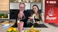 Andy Ogris und Tanja lächeln gemeinsam in einer modernen Küche hinter einer Küchentheke. Andy hält ein hohes Glas mit rosa Nicecream hoch, während Tanja eine Erdbeere auf ihrer Handfläche präsentiert. Im Vordergrund auf der Arbeitsfläche sind ein weiteres serviertes Glas Nicecream sowie Zutaten wie Bananen, Zitrusfrüchte und ein Holzbrett mit Cookie-Dough zu sehen.