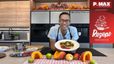 Chefkoch Peter Lehner präsentiert lächelnd einen Teller mit dem zubereiteten Carpaccio in einer modernen Showküche. Im Vordergrund sind herbstliche Zutaten wie halbierte Kürbisse, Äpfel und Limetten auf der Arbeitsplatte angerichtet.
