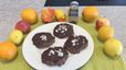 Vier runde Elisenlebkuchen mit dunklem Schokoladenüberzug und Mandelblättchen liegen auf einem weißen Teller. Der Teller ist dekorativ umrahmt von frischen Orangen, Zitronen, Äpfeln und einem Gewürzstreuer mit der Aufschrift „Lebkuchen“.