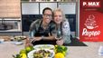 Peter Lehner und Schauspielerin Susanna Hirschler stehen lächelnd in einer modernen Küche nebeneinander und halten jeweils ein Glas Weißwein. Vor ihnen auf dem Küchentresen sind fertig angerichtete mediterrane Gerichte mit Seeteufel und Oktopus sowie Zitronen und Limetten zu sehen.