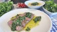 Tranchiertes, rosa gegartes Steak auf einem weißen Teller, garniert mit frischer, grüner Chimichurri-Sauce und Olivenöl. Im Hintergrund sind Schüsseln mit Macaroni-Mais-Gratin und frischen Kräutern angerichtet.