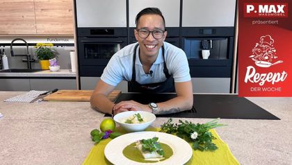 Chefkoch Peter Lehner lächelt in einer modernen Küche hinter dem fertig angerichteten Gericht: gebratener Kabeljau auf einem Spiegel aus grüner Currysauce sowie eine Schale Jasminreis, dekorativ angerichtet mit frischen Kräutern und einer Limette.