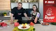 Schauspieler Max Ortner und Moderatorin Tanja stehen in einer modernen Küche hinter einer Kochinsel und präsentieren den fertig zubereiteten Wassermelonen-Feta-Salat mit einem Parmesan-Chip. Auf der Arbeitsplatte im Vordergrund ist der angerichtete Teller zu sehen, umgeben von frischen Wassermelonenstücken und Limetten.
