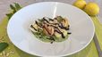 Angerichtet auf einem tiefen, weißen Teller: Selbstgemachte Tortellini auf einer hellgrünen Sauce, garniert mit gebratenen Garnelen, dunkler Balsamico-Creme und frischen Minzblättern. Im Hintergrund sind Zitronen, Pinienkerne und Minze auf einer hellgrünen Unterlage zu sehen.