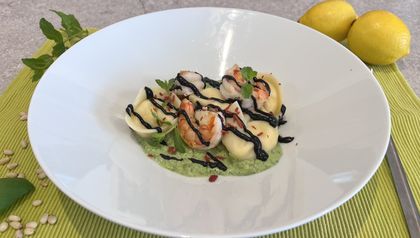 Angerichtet auf einem tiefen, weißen Teller: Selbstgemachte Tortellini auf einer hellgrünen Sauce, garniert mit gebratenen Garnelen, dunkler Balsamico-Creme und frischen Minzblättern. Im Hintergrund sind Zitronen, Pinienkerne und Minze auf einer hellgrünen Unterlage zu sehen.
