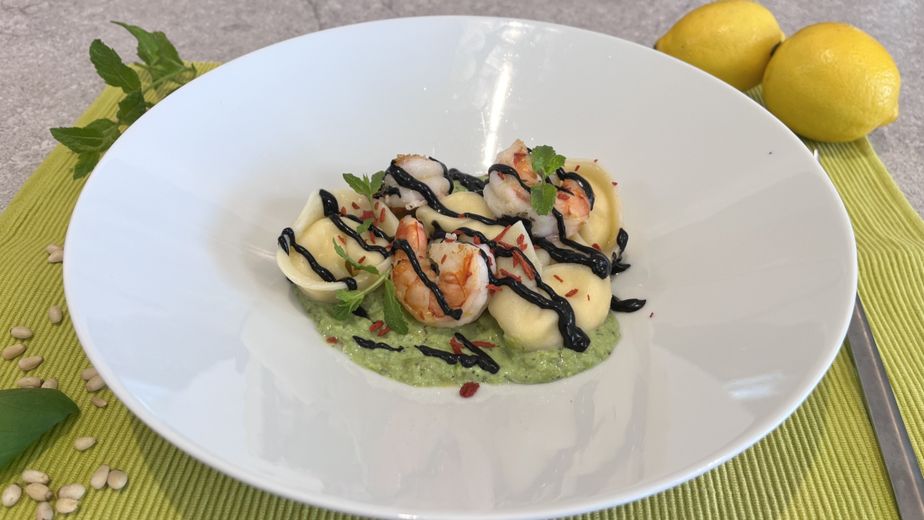 Angerichtet auf einem tiefen, weißen Teller: Selbstgemachte Tortellini auf einer hellgrünen Sauce, garniert mit gebratenen Garnelen, dunkler Balsamico-Creme und frischen Minzblättern. Im Hintergrund sind Zitronen, Pinienkerne und Minze auf einer hellgrünen Unterlage zu sehen.