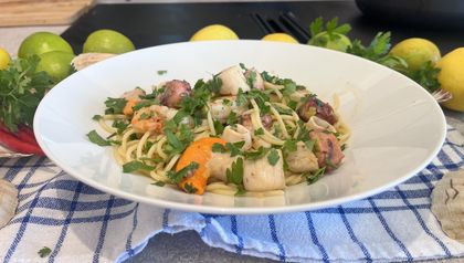 Ein tiefer weißer Teller mit frisch zubereiteter mediterraner Seafood-Pasta, angerichtet auf einem blau-weiß karierten Geschirrtuch. Auf den Spaghetti liegen Oktopusstücke, Garnelen, Jakobsmuscheln und Calamari-Ringe, garniert mit reichlich gehackter Petersilie.
