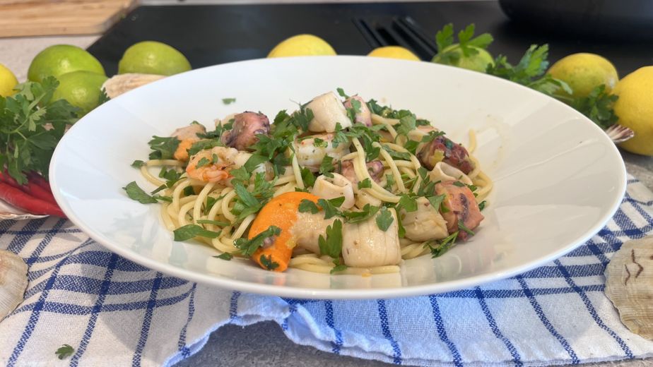 Ein tiefer weißer Teller mit frisch zubereiteter mediterraner Seafood-Pasta, angerichtet auf einem blau-weiß karierten Geschirrtuch. Auf den Spaghetti liegen Oktopusstücke, Garnelen, Jakobsmuscheln und Calamari-Ringe, garniert mit reichlich gehackter Petersilie.