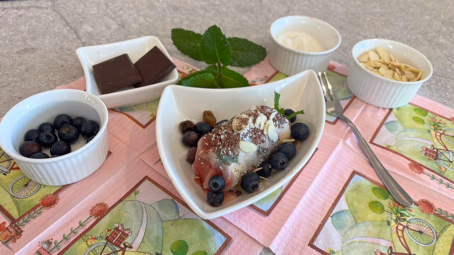 Eine fertig zubereitete süße Sommerrolle, angerichtet in einer geschwungenen weißen Schale und garniert mit Heidelbeeren, Schokoladenraspeln und Mandelblättchen. Ringsherum stehen kleine Schälchen mit den Zutaten wie Heidelbeeren, dunkler Schokolade, Kokosraspeln, Mandelblättchen sowie frischer Minze.