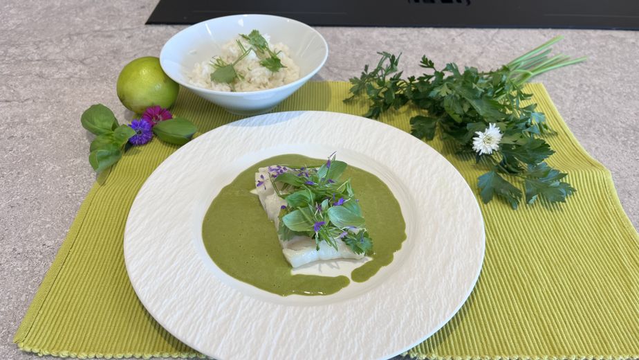 Gebratener Kabeljau, serviert in einer cremigen, hellgrünen Currysauce und garniert mit frischen Kräutern sowie essbaren lila Blüten auf einem weißen Teller. Im Hintergrund ist eine Schale mit Jasminreis, frischen Kräutern und einer Limette auf einem hellgrünen Tischset angerichtet.