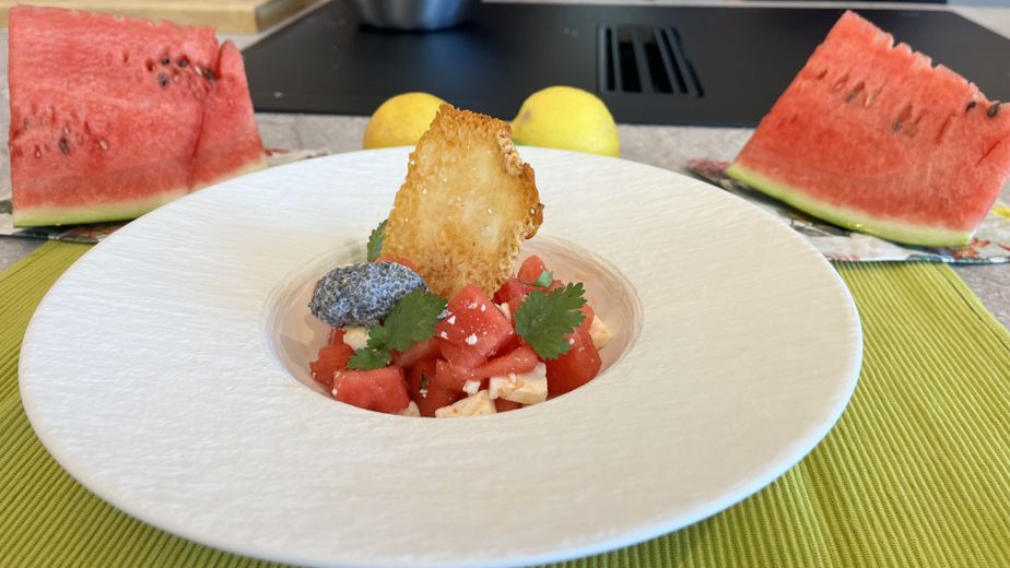 Ein elegant angerichteter Wassermelonen-Feta-Salat in einem tiefen, weißen Teller mit breitem Rand. Der Salat mit frischen Kräutern wird von einem großen, knusprigen Parmesan-Chip gekrönt, während im Hintergrund frische Wassermelonenstücke und Zitronen dekorativ platziert sind.