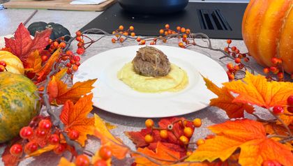 Ein weißer Teller mit cremigen Erdäpfel-Lauch-Püree, auf dem ein faschiertes Laibchen mit geschmorten Zwiebeln angerichtet ist. Der Teller steht auf einer Küchenarbeitsplatte, umrahmt von herbstlicher Halloween-Dekoration aus buntem Laub, Kürbissen und Beerenranken.