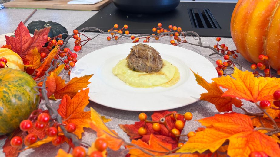 Ein weißer Teller mit cremigen Erdäpfel-Lauch-Püree, auf dem ein faschiertes Laibchen mit geschmorten Zwiebeln angerichtet ist. Der Teller steht auf einer Küchenarbeitsplatte, umrahmt von herbstlicher Halloween-Dekoration aus buntem Laub, Kürbissen und Beerenranken.