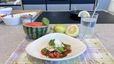 Ein fertig angerichteter Wassermelonensalat mit Burrata, frischem Basilikum und Pinienkernen in einem weißen, geschwungenen Teller. Im Hintergrund stehen eine halbe Wassermelone, Zitronen und ein Glas Wasser mit einer Zitronenscheibe und Strohhalm auf einer Küchenarbeitsplatte.