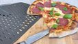 Eine frisch gebackene Salami-Pizza mit geschmolzenem Käse und frischem Basilikum liegt auf einem Holzschneidebrett, von dem bereits ein Stück abgeschnitten wurde. Daneben sind ein großes Küchenmesser und ein perforierter schwarzer Pizzaschieber zu sehen. Der dicke, luftige Rand der Pizza zeigt eine knusprige, leicht gebräunte Backkruste.