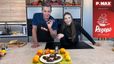 Tanja und Philipp stehen lächelnd in einer modernen Küche an einer Kochinsel. Vor ihnen auf der Arbeitsplatte ist ein Teller mit frisch zubereiteten Elisenlebkuchen angerichtet, dekoriert mit Orangen, Zitronen und herbstlichen Blättern.