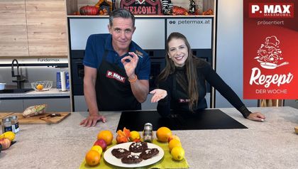 Tanja und Philipp stehen lächelnd in einer modernen Küche an einer Kochinsel. Vor ihnen auf der Arbeitsplatte ist ein Teller mit frisch zubereiteten Elisenlebkuchen angerichtet, dekoriert mit Orangen, Zitronen und herbstlichen Blättern.