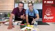Philipp Jelinek und Chefkoch Peter Lehner stehen lächelnd in einer Küche hinter einer Arbeitsplatte, auf der ein fertig angerichteter Wassermelonensalat mit Burrata präsentiert wird. Vor ihnen liegen zudem eine halbe Wassermelone, Zitronen und ein Glas Wasser.