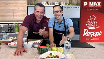 Philipp Jelinek und Chefkoch Peter Lehner stehen lächelnd in einer Küche hinter einer Arbeitsplatte, auf der ein fertig angerichteter Wassermelonensalat mit Burrata präsentiert wird. Vor ihnen liegen zudem eine halbe Wassermelone, Zitronen und ein Glas Wasser.